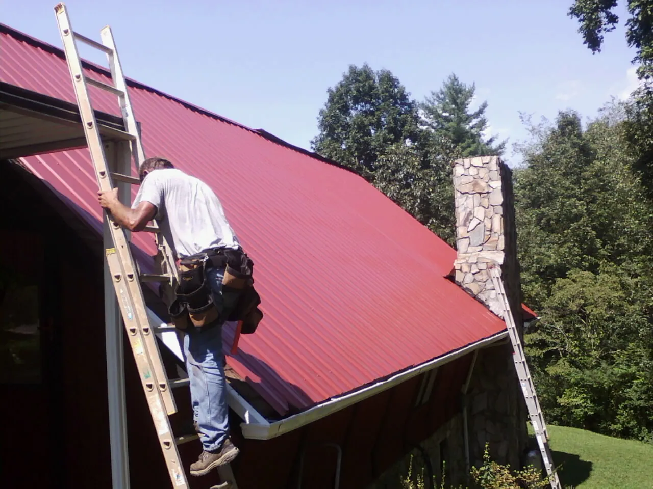 Roofer on a ladder beside a red metal roof and stone chimney — Soffit & Fascia Repair by Roofing Brothers in South Yarmouth